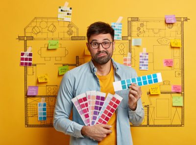 Hesitant bearded man painter chooses color from samples, decides to refurbish rooms in house, works on design, stands against drawn sketch with sticky notes and palette. Repair, building concept