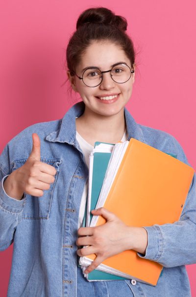 Excited young student wearing denim jacket and eyeglasses, holding colorful folders and showing thumb up, looking directly at camera, dark haired female with hair bun expressing positive emotions.